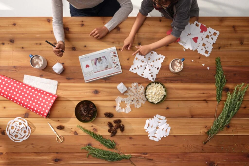 View from above of two people making paper snowflakes on a large wooden table