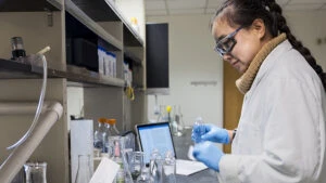 A researcher wearing lab attire is working in a laboratory packed with glass beakers and a laptop.