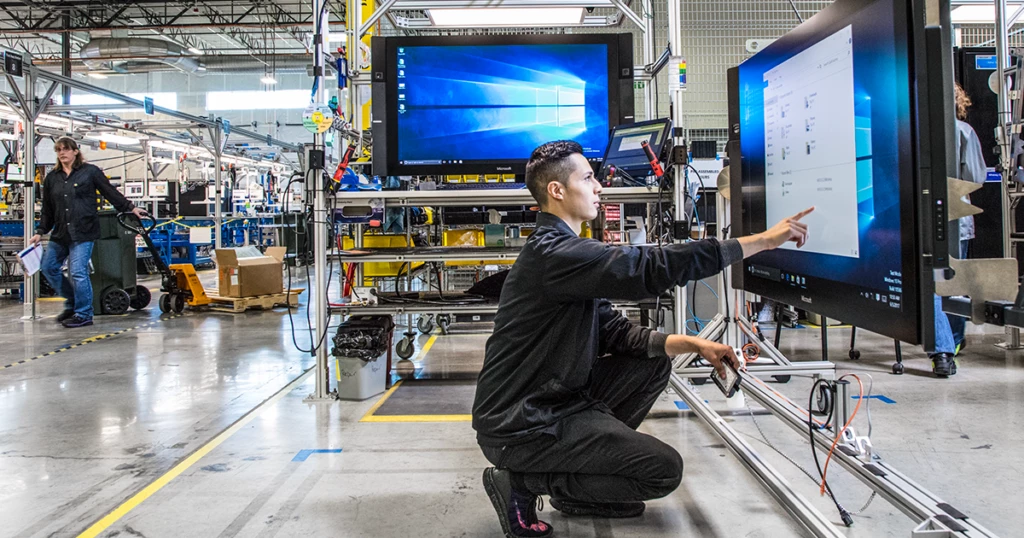 A millennial man crouches down to use a large touchscreen monitor in a test factory.  Another large monitor and a female worker using a hand truck are shown in the background.