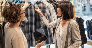 Female store employee using tablet standing next to female consumer in front of retail clothing display. The employee is checking the price tag.