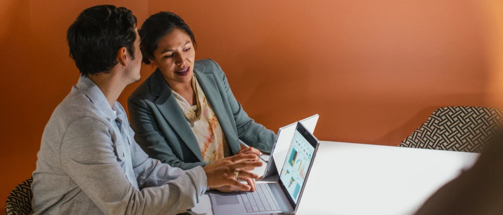 Two colleagues working on laptops side by side in a boardroom.