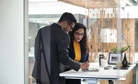 Two employees collaborate over a tablet at a desk in an office setting.