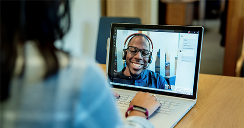 Image of a woman at a desk using a Surface laptop to make a Microsoft Teams video call with one man smiling and wearing a headset.