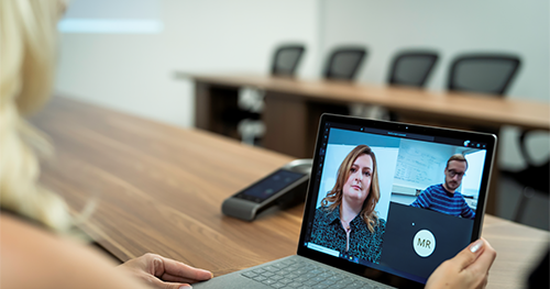 An image of a blond haired woman in an empty conference room using a Surface laptop to make a Microsoft Teams video call with three other people.