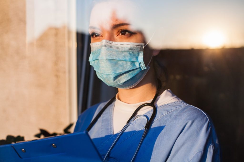 A woman standing in a window, wearing nurses scrubs and a PPE mask., holding a clip board.