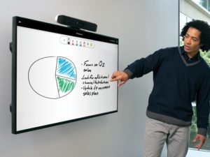 One male pointing to a wall-mounted display featuring whiteboard functinality with a Poly Microsoft Teams Rooms on Android collaboration bar wall on top of the display
