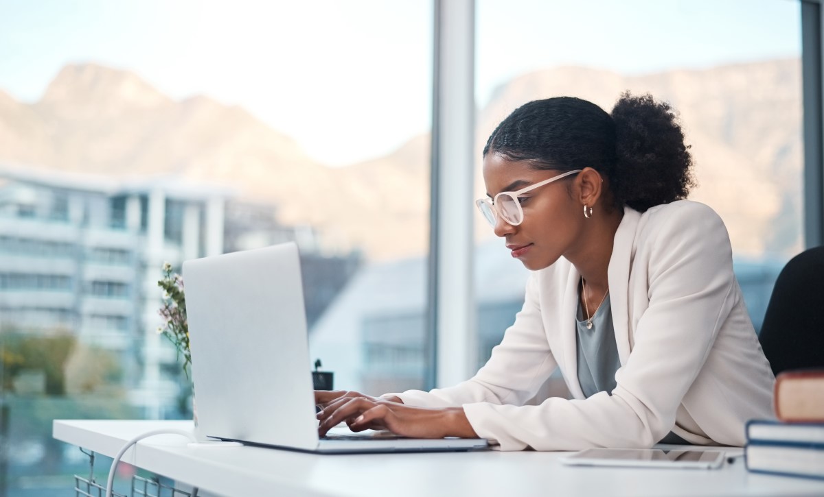 Picture of a woman wearing a white blazer and glasses, looking down at a laptop on her desk.
