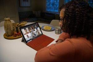 Over the shoulder photo of an adult female in kitchen participating in a Microsoft Teams video call on a Surface Pro with Poppy Red type cover.