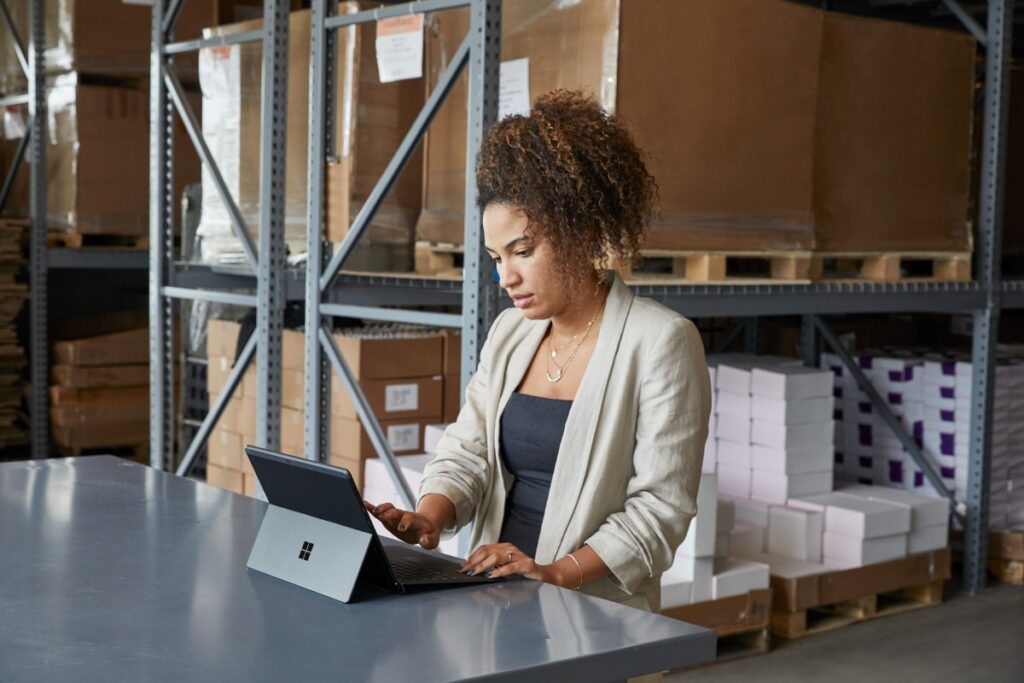 Adult female in a warehouse setting, sitting at a table using a platinum Microsoft Surface Pro X in laptop mode.