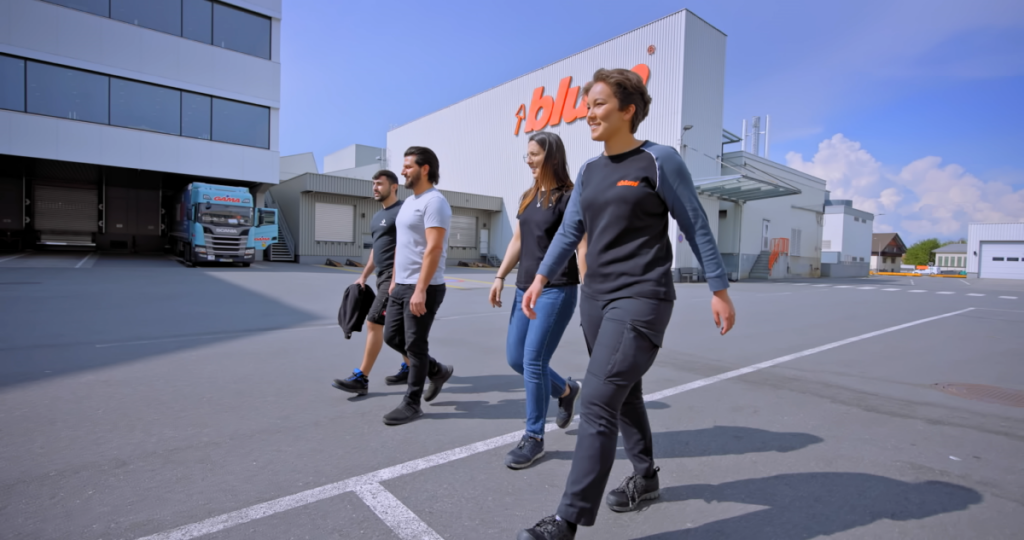 Video still of happy employees walking outside of Blum manufacturing building with company logo.