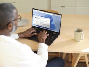 Man sitting at a wooden table typing on a laptop while using Microsoft Teams within Windows 365. Shown on Windows 11. Keywords: Markus Long; Fabrikam; one screen; view over the shoulder; Edge browser
