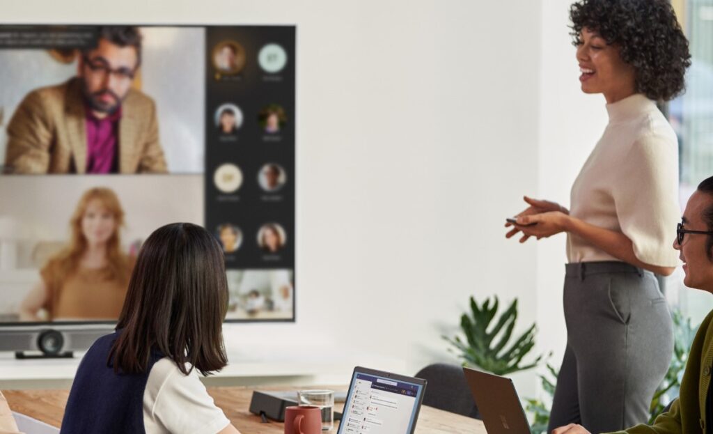 A female speaker presenting in a Microsoft Teams Room (MTR) meeting while people in the conference room are viewing remote attendees in Gallery Mode with participant chat on the front of room (FoR) screen​. Keywords​: Logitech Rally Bar Mini; Logitech Tap console; male; man; women; colleagues; woman is chatting on MS Teams on her laptop; collaboration; collaborating