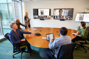 A coordinated meeting is taking place in a Microsoft Teams Room; two women, three men (one man in a wheelchair) are meeting with several team members joining remotely in Front of Room view. The in-person team is sharing a PowerPoint presentation from a Surface laptop, casting onto a Surface Hub and the Teams Room, allowing more opportunity for collaboration.