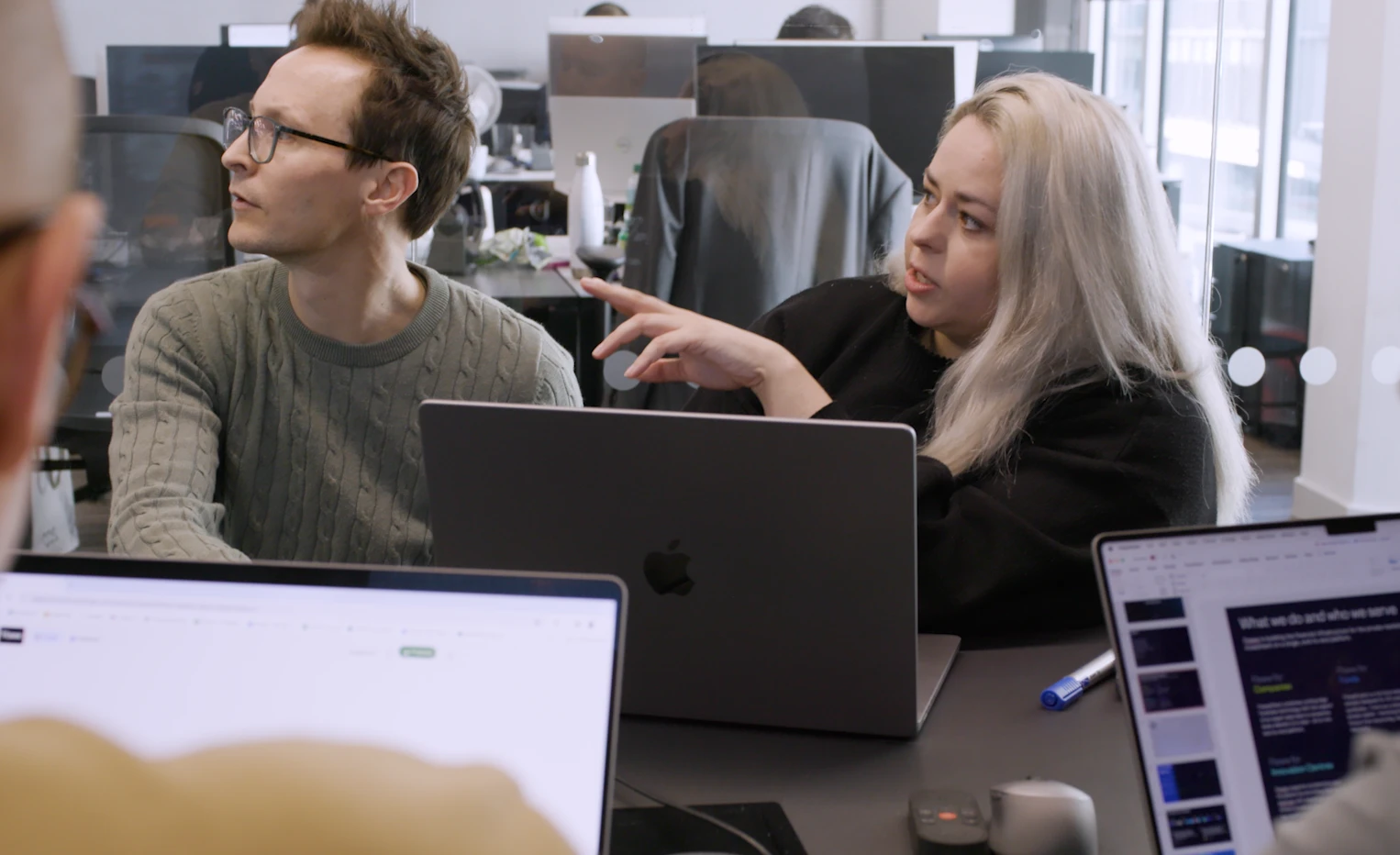 A man and a woman sitting at a table with laptops looking at a screen off camera. The woman is talking.