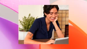 A person sitting at a table, smiling, working on a laptop. The photo is overlaid on a magenta and orange graphic design background.