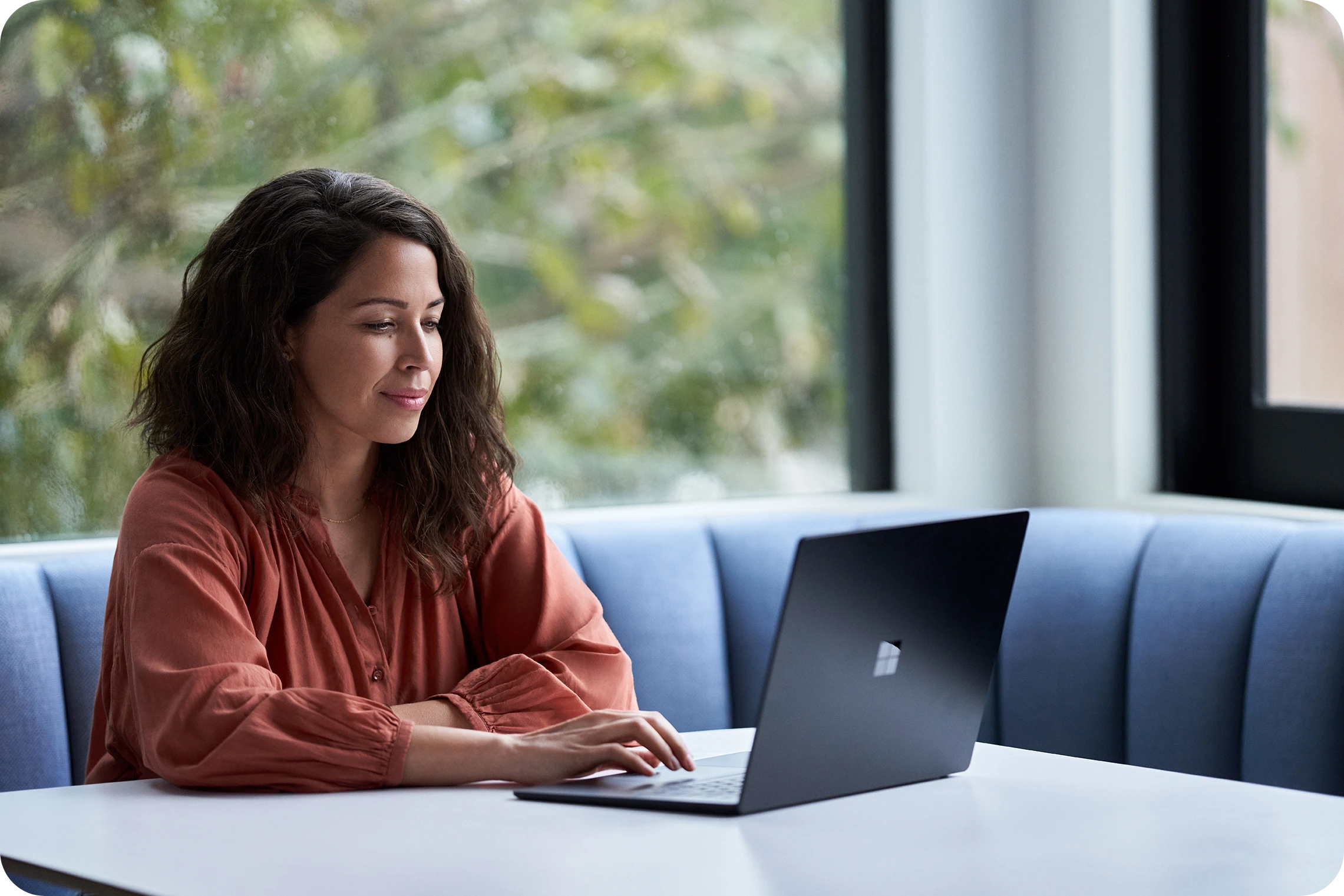 Person working on a Microsoft Surface laptop at a table in a bright, modern workspace with large windows.