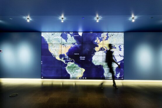A blurry person walking in front of a large map of the globe on a large screen.