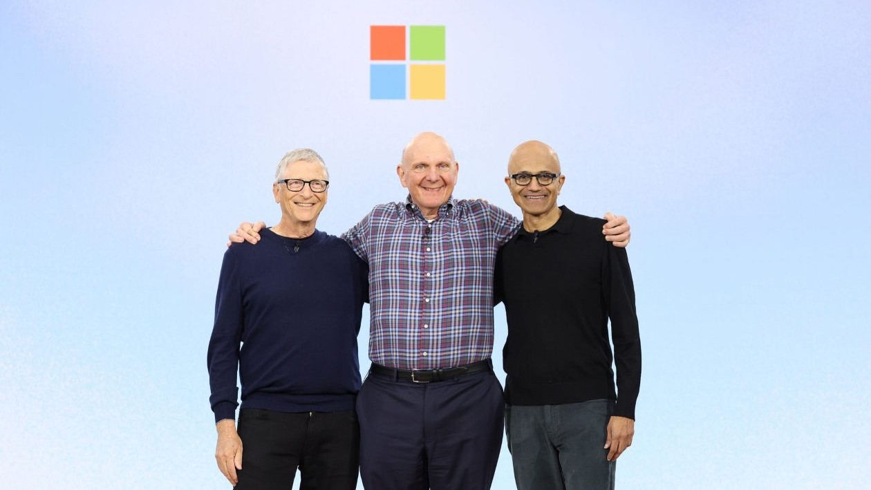 Three men with their arms around each other’s shoulders stand on stage in front of the colorful square Microsoft logo.
