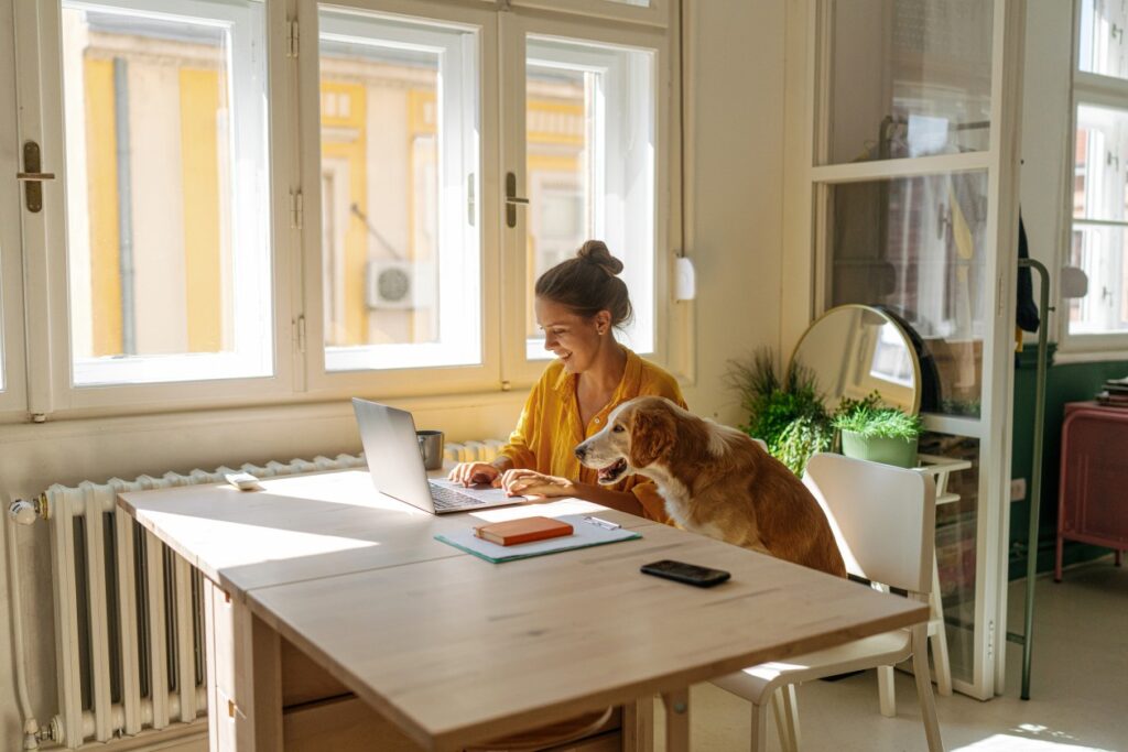 Woman working from her home office. She is using a laptop and her pet dog is sitting a chair next to her.