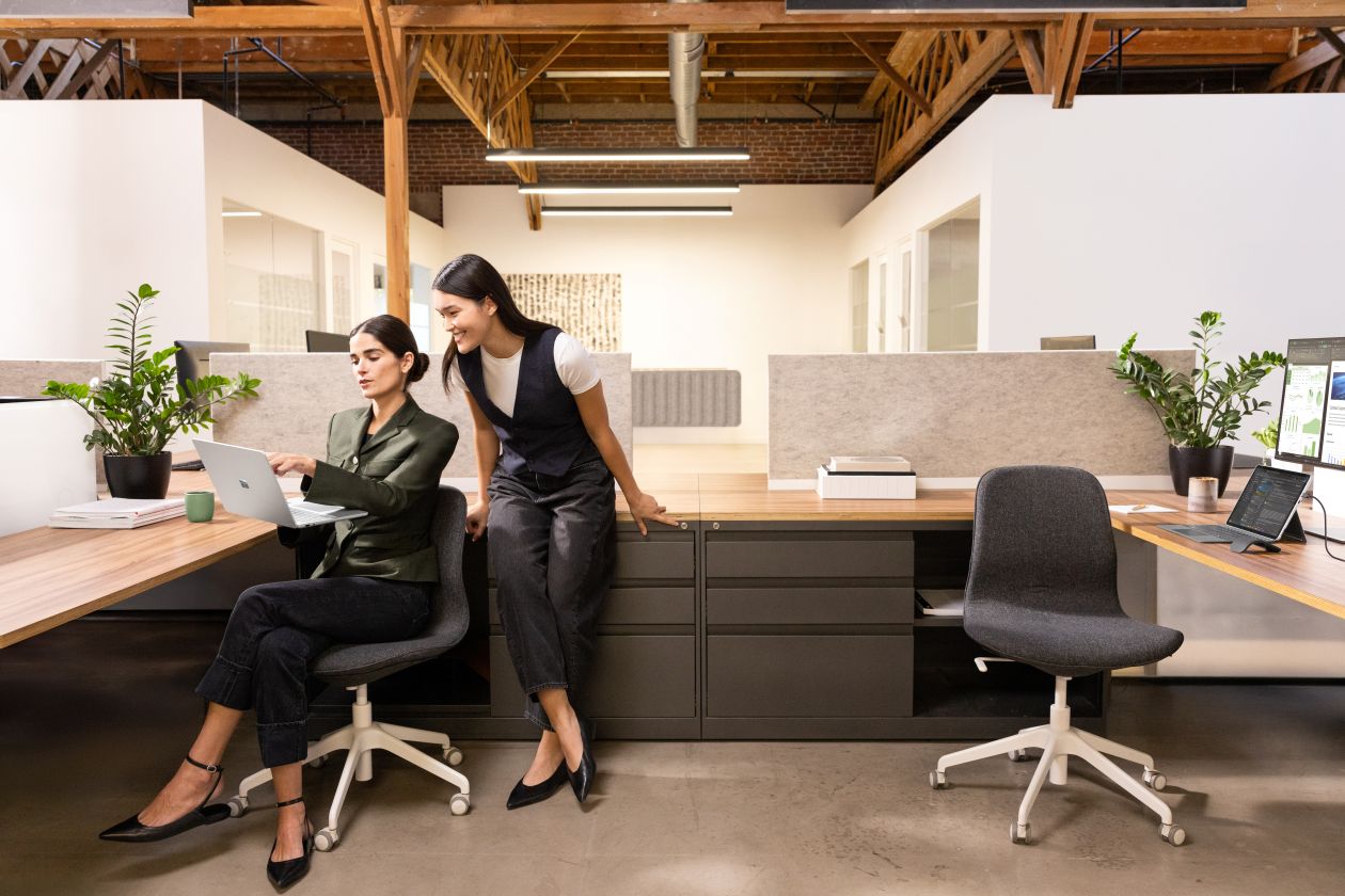 Photograph of two female executives in an office. One executive is seated, holding a Surface Laptop 13" Platinum device in her right hand. A Surface Pro 12" Platinum device in laptop mode is on another desk, connected to an external monitor. A Surface Arc Mouse is next to the device.