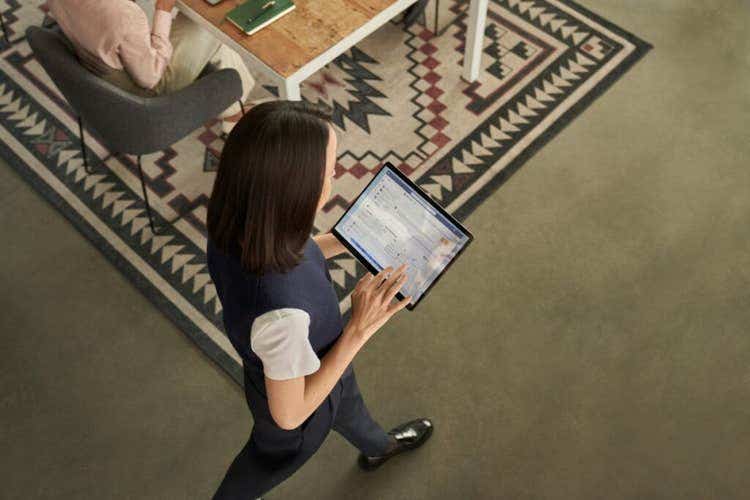 Woman walking across an office floor looks down at a tablet she is using, while another person works at a wooden table with a patterned rug in the background
