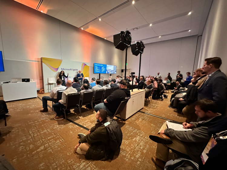 Audience seated and listening to a panel discussion at the Microsoft Ignite event, inside a conference room with stage lighting and a ‘Microsoft Ignite’ backdrop. The room is filled with attendees in chairs and on the floor, with presenters speaking at the front.