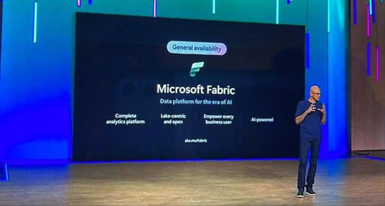 A speaker on stage at a major enterprise-tech event presenting the ‘General Availability’ announcement of Microsoft Fabric. The large screen behind shows the Microsoft Fabric logo and key phrases such as ‘Data platform for the era of AI’, ‘complete analytics platform’, ‘lake-centric and open’, and ‘AI-powered’. The presenter gestures toward the screen in a dark auditorium with stage lighting.
