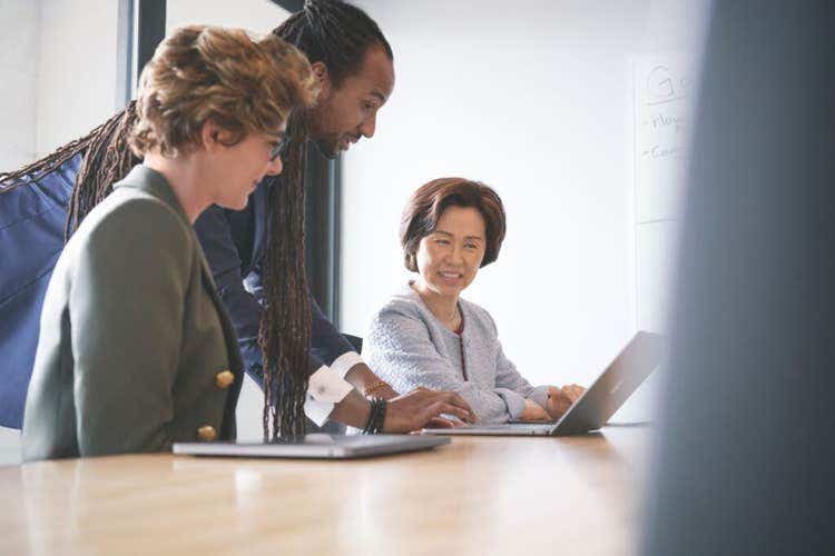 Three colleagues gather around a conference table, with one person seated at a laptop while the other two stand and lean in, smiling and collaborating on the screen’s content