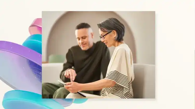 Two colleagues sit together on a light-colored couch in a softly lit room, smiling and discussing something on a tablet