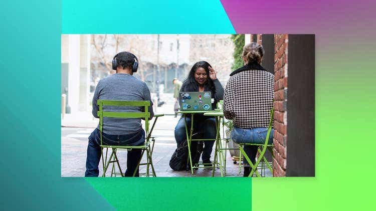 Three people work at bright green café tables in an outdoor alleyway, with the person in the center smiling at a sticker-covered laptop, all framed by a teal, purple, and lime-green gradient background