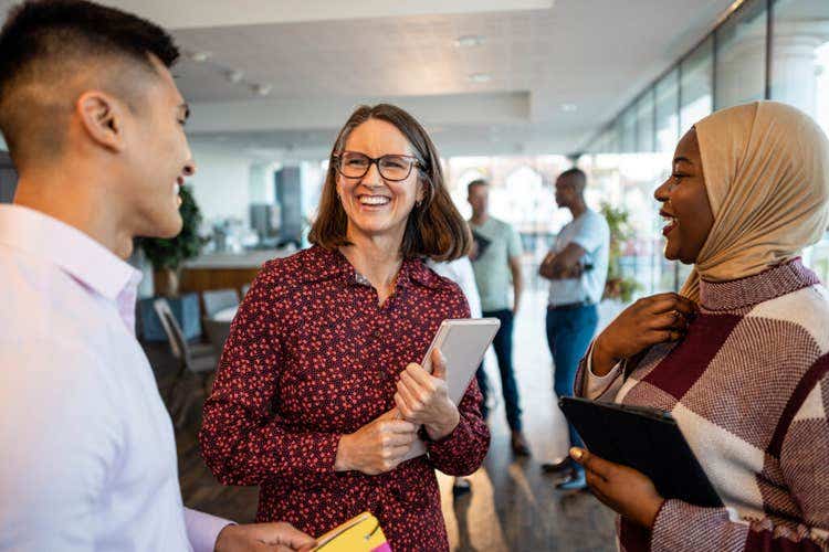 Group of colleagues stand and chat in a modern office, with a woman holding a tablet and smiling at two coworkers who face her while others talk in the background
