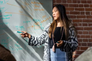 Real people, real offices. Female developer speaking in front of a white board during team stand up meeting. Women who code, women developers, women engineers, code, develop, developer, engineer, Visual Studio, Azure.