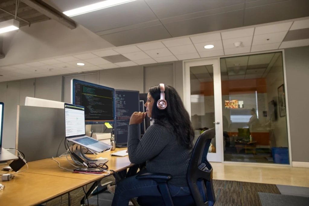 Real people, real offices. Black female developer wearing headphones, coding at her desk.
