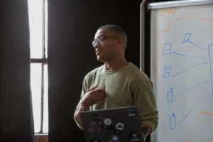 Real people, real offices. Male developer speaking in front of a white board during team stand up meeting, holding a Surface laptop personalized with stickers. Code, develop, developer, engineer, Visual Studio, Azure.