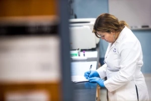 Female technician wearing lab coat and gloves writes with pen and paper in medical laboratory of Chicago hospital.