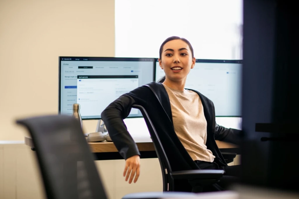 Engineer working at her desk; multi-screen workstation. She turns to face a colleague to discuss the current project.