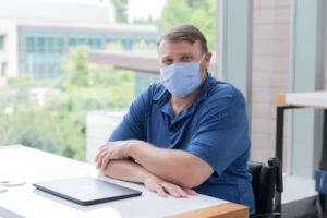 A male employee in a wheelchair wearing a face mask and smiling at his desk. Hybrid Workplace collection.