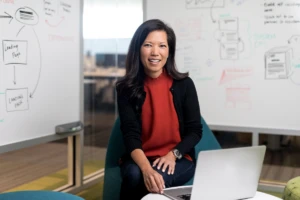 Portrait of female executive in red shirt, smiling and facing camera. She is in an office conference room setting, using an open laptop (screen not shown). Two whiteboards are partially visible in the background, with diagrams and flowcharts written on them.