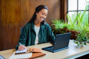 Woman sitting at a casual desk at home with an HP Elite Folio, taking notes and smiling.