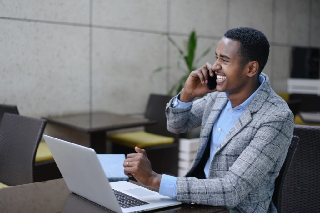 Portrait of a young man working on his laptop smiling on his cellphone