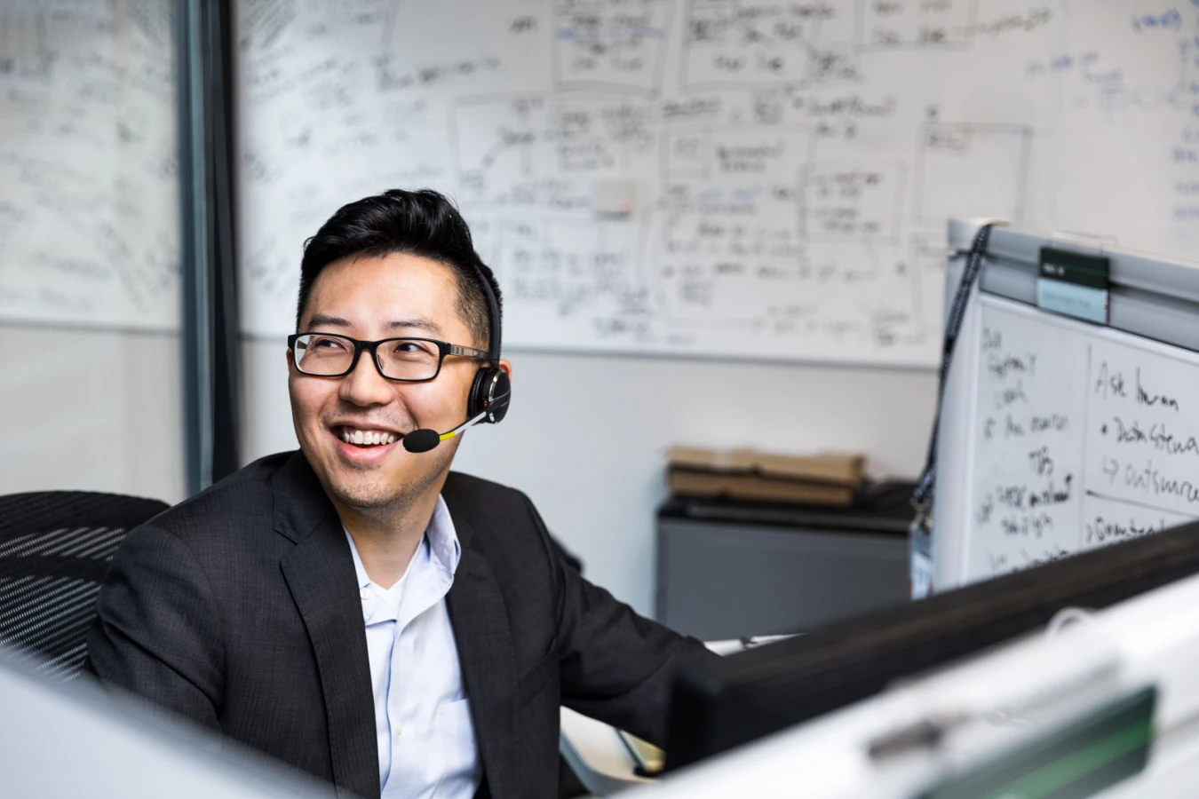 Male worker sitting at cubicle desk in financial office, wearing glasses and phone headset. He is smiling and looking up to his right. A large whiteboard covered in black written text is on wall behind him. A smaller whiteboard also partially visible on his desk&lsquo;s cubicle wall.