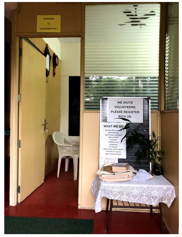 A view of a small room, where helpline calls are taken by volunteers, from just outside the door. A chair sits in the room, and just outside the door is a table with a plant and a side that reads 