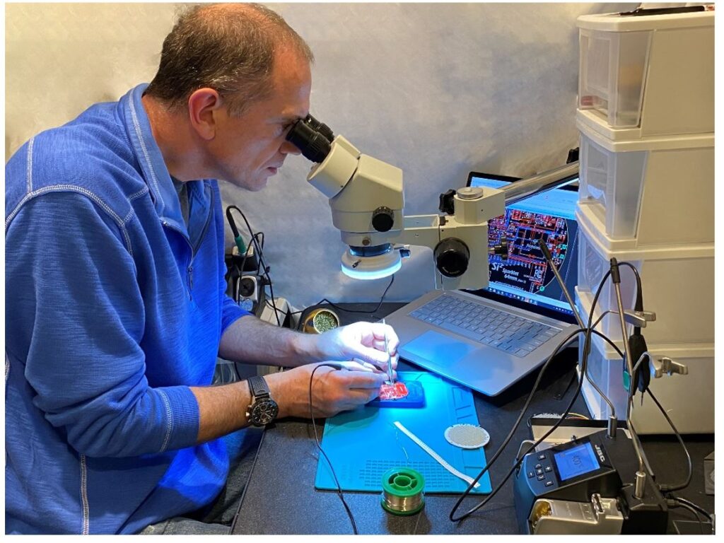 A man peers through a microscope while soldering a small red motherboard. The view from the microscope is displayed on a laptop on the desk next to the microchip.