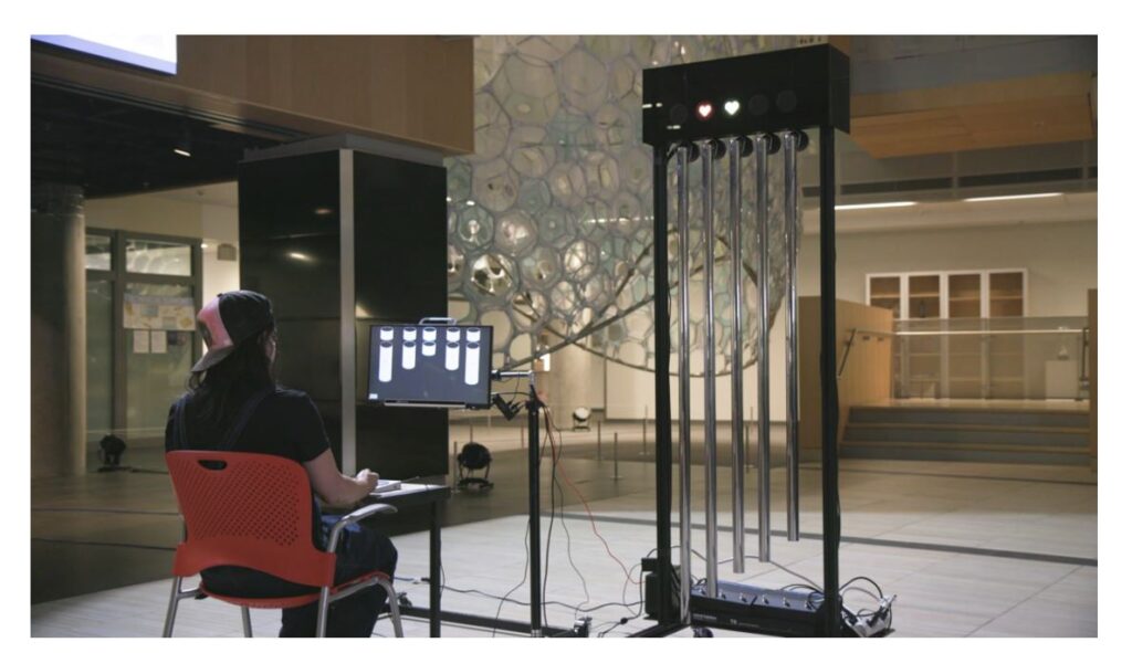 A woman sits in front of a computer screen and controls a set of tubular bells using a control interface on a table. 