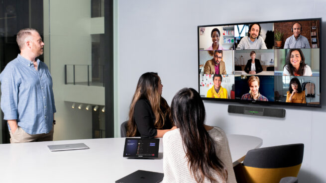 One male and two females in medium conference room with a Crestron Microsoft Teams Rooms on Android device and touch display being used for remote video meeting with Teams Meetings. Large mounted display showing remote participants and two Surface Device