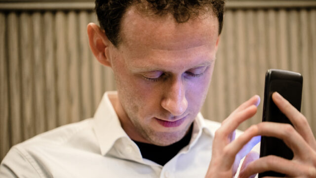 A close-up of a man wearing casual clothing, he has his smartphone in his hand and he is using an assistive mobile app for people with vision disabilities to assist him.
