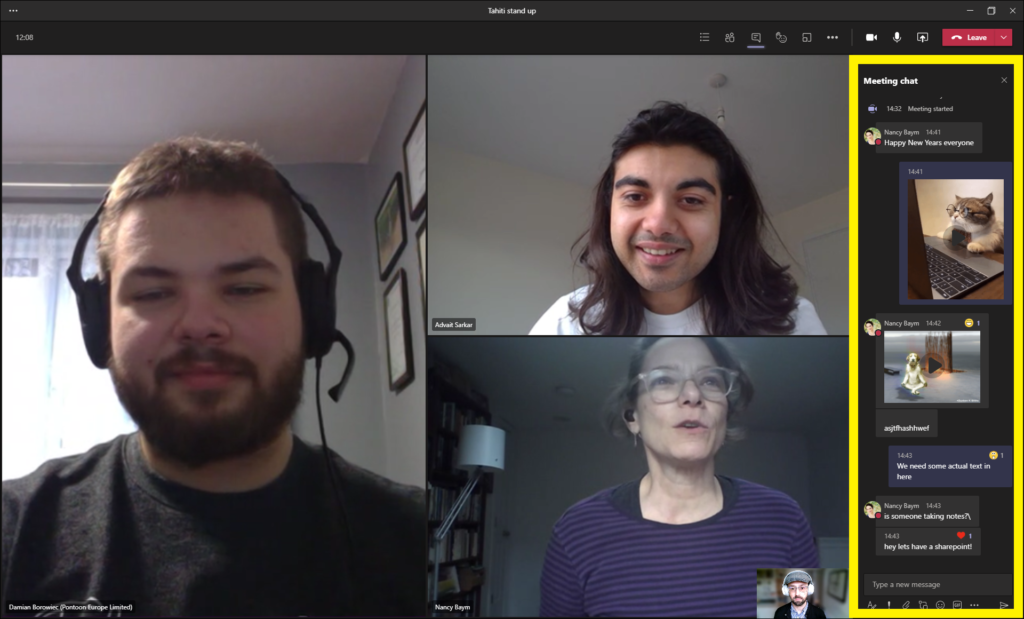 Three people meet in Microsoft Teams. On the left, a person with a beard is wearing headphones with a microphone and is smiling. They wear a black t-shirt, and they are standing in front of a curtained window in a house. A wall to his left has a gallery of pictures. Moving clockwise, another person has long, dark hair and is also smiling. They wear a white t-shirt. They are standing in a room with white walls, and a turned-off lightbulb hangs above their head. Next, a person is talking. They have glasses, a purple shirt on with horizontal stripes, and stand in a room with a bookshelf and floor lamp to their right. There is also another person in a small window at the bottom of the screen, wearing a hat, cardigan, and headphones. The sidebar chat is outlined with a yellow border. From top to bottom, the chat window reads: Meeting Chat. Nancy Baym: “Happy New Years everyone.” A picture of a cat with glasses working at a laptop. Nancy Baym: A video of a dog levitating next to a tree. This video has a “laugh” reaction. A string of unintelligible text in the chat follows. “We need some actual text in here.” Nancy Baym: “Is someone taking notes? Hey let’s have a sharepoint!” A heart reaction shows on the last comment.