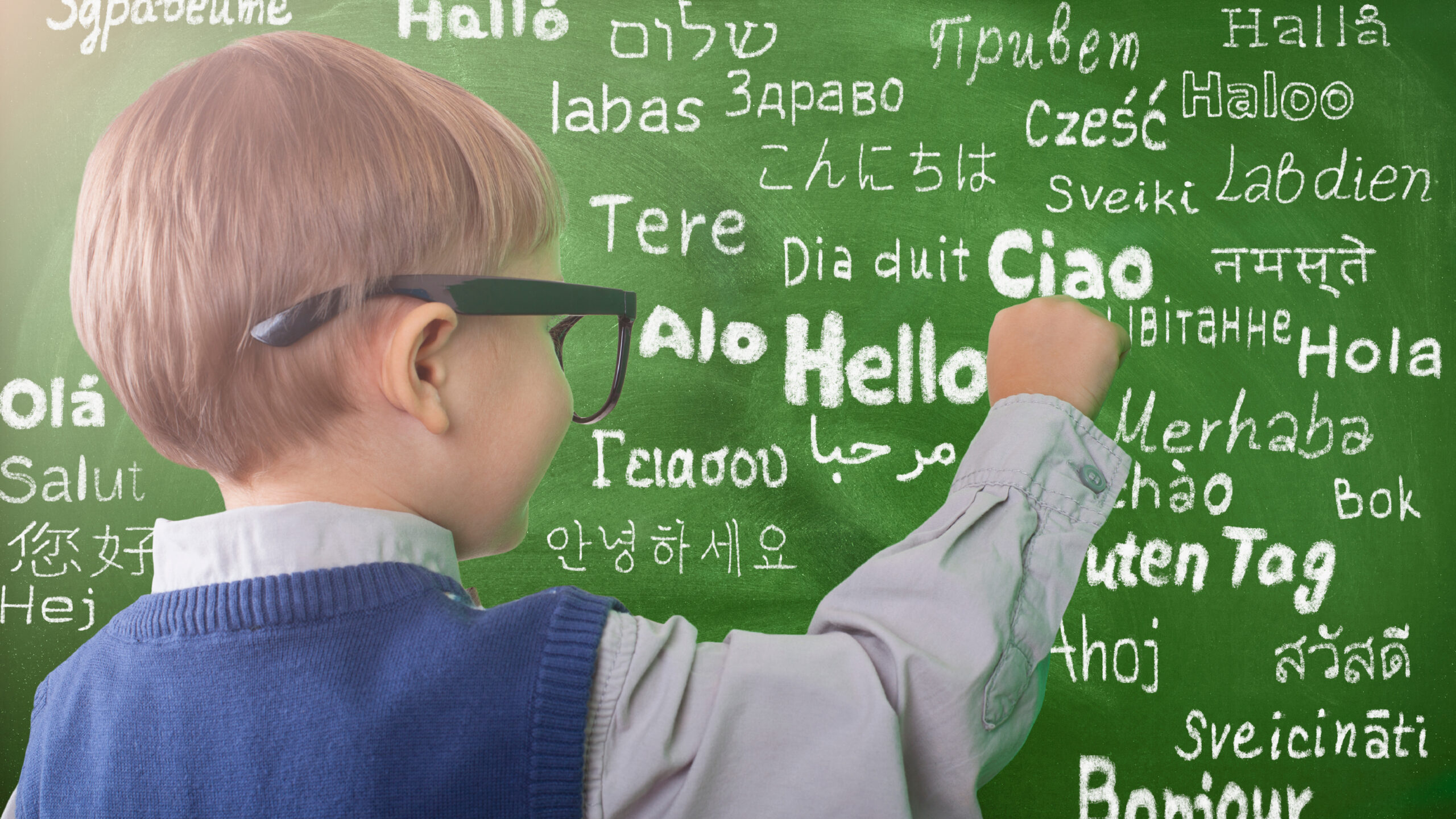 Photo of a young child writing on a chalkboard. The word “hello” is written on the chalkboard in multiple languages.