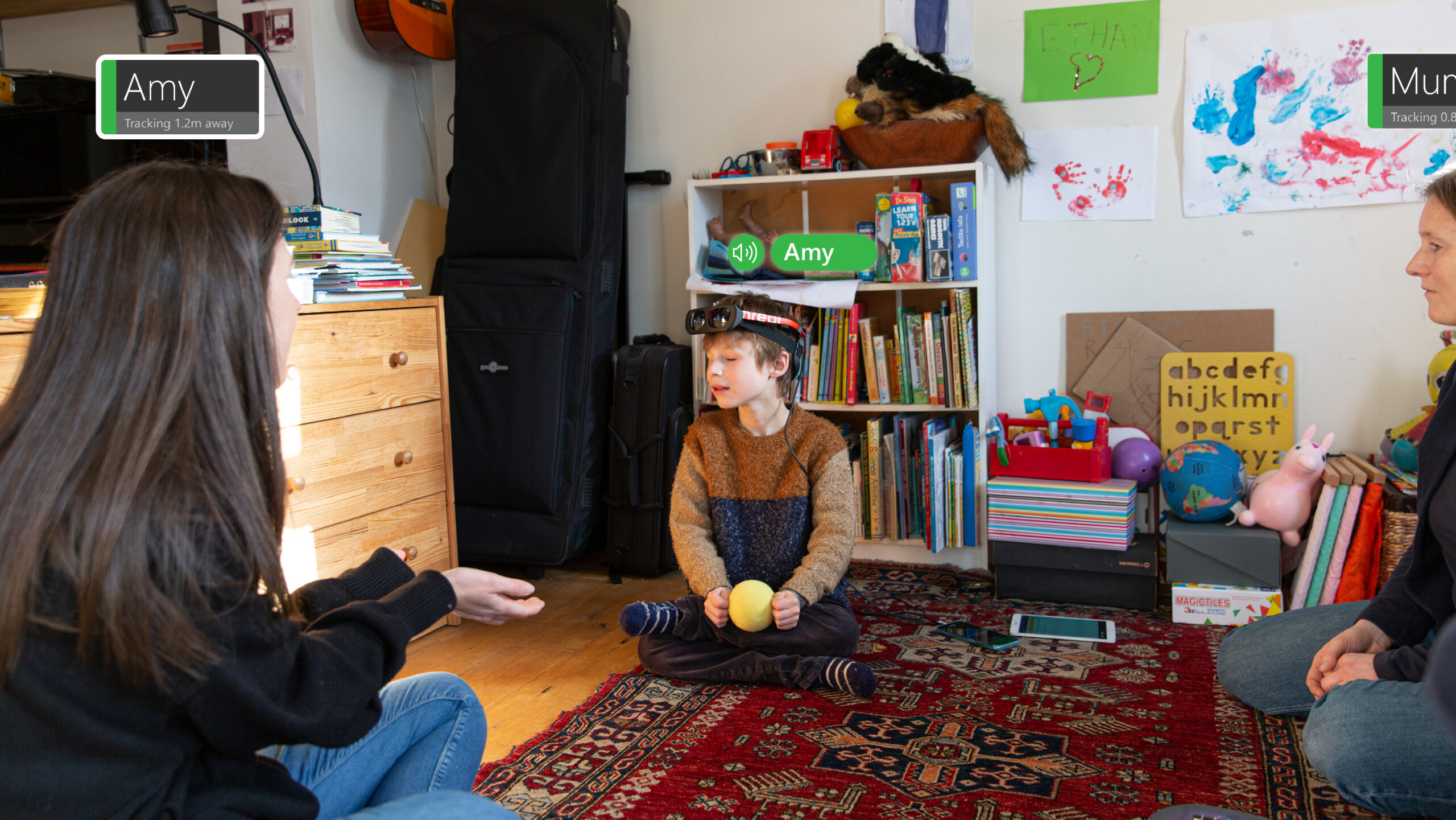A young boy wearing the PeopleLens sits on the floor of a playroom holding a blind tennis ball in his hands. His attention is directed toward a woman sitting on the floor in front of him holding her hands out. The PeopleLens looks like small goggles that sit on the forehead. The image is marked with visual annotations to indicate what the PeopleLens is seeing and what sounds are being heard.