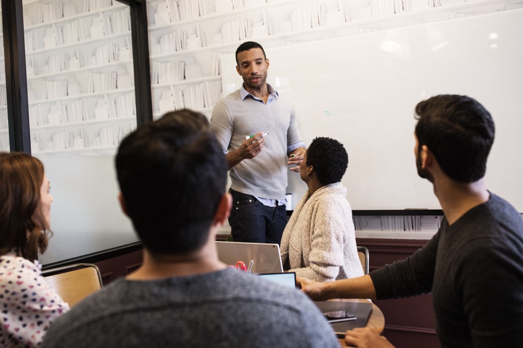 Photograph of a group of people in an office like setting. One person stands in front of a whiteboard.